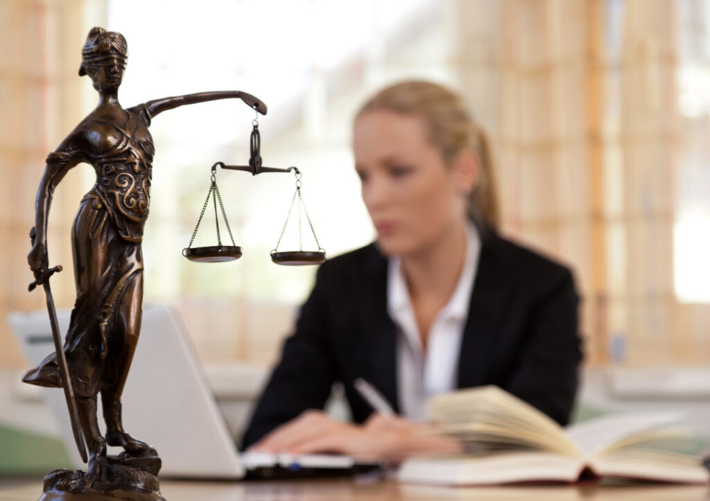 Eine junge Rechtsanwältin sitzt an ihrem Schreibtisch im Hintergrund, während die linke Seite von einer Statue von Justizia scharf im Vordergrund zeigt.